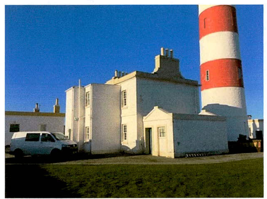 A photograph showing a white building complex situated next to a tall red and white striped lighthouse under a clear blue sky.