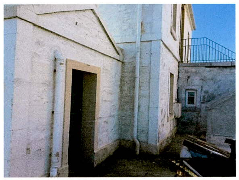Exterior photograph of a white-washed stone building featuring a doorway, drainpipe, and a balcony railing in the background.