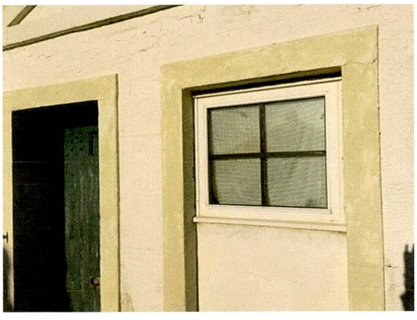 A close-up photograph of a cream-colored building exterior featuring a white four-pane window and a dark doorway, both framed by light stone surrounds.