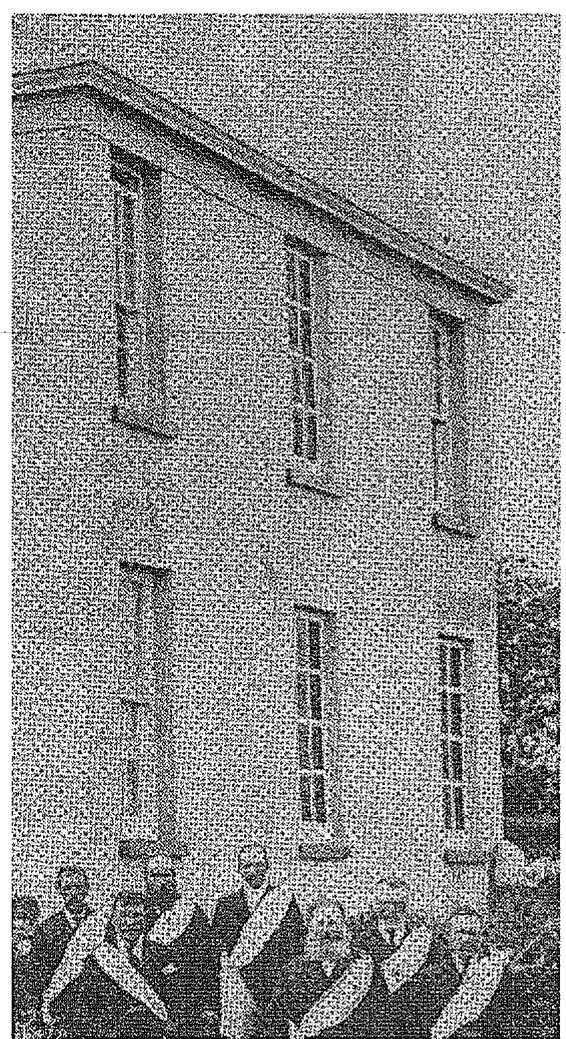 A grainy, black and white archival photograph showing the exterior facade of a large two-story building with a group of people standing in front.