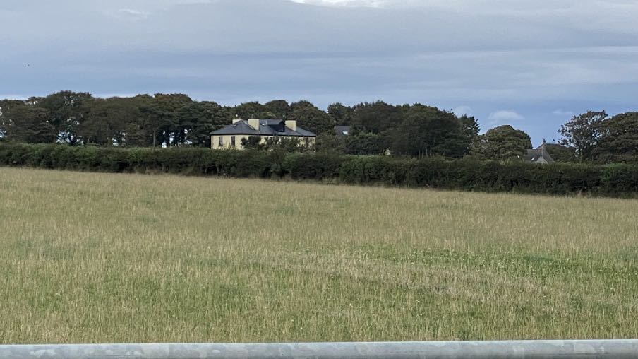 A photograph showing a large detached house, Ballaradcliffe House, situated behind a dense hedge and trees, viewed from across a grassy field.