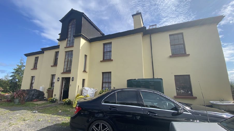 A low-angle photograph of a large, cream-colored detached house with a distinctive gabled roof section and a black car parked in the foreground.