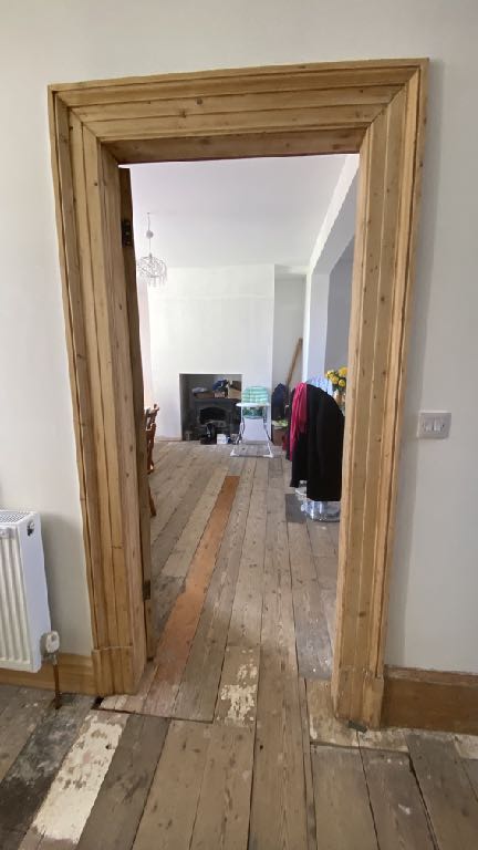 Interior photograph showing a large rustic wooden door frame leading into a room with wooden floorboards and a fireplace.