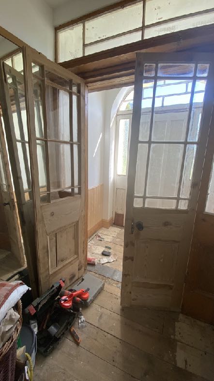 Interior photograph of a room with wooden floors and large glass-paned doors, showing renovation tools and materials on the floor.