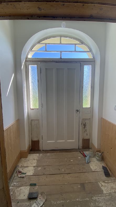 An interior photograph showing a white entrance door with an arched fanlight and side panels, set within a hallway featuring wooden floorboards and wainscoting.