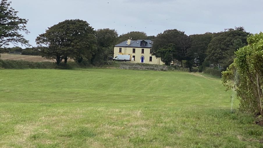 A photograph showing a large, two-story cream-colored house situated on a slight rise, surrounded by trees and a large grassy field in the foreground.
