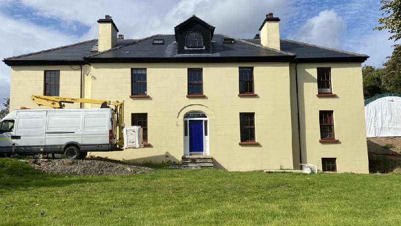 A large, two-story yellow detached house with a black roof and blue door is shown in a grassy setting. A white van with a yellow cherry picker is parked on the left side of the property.