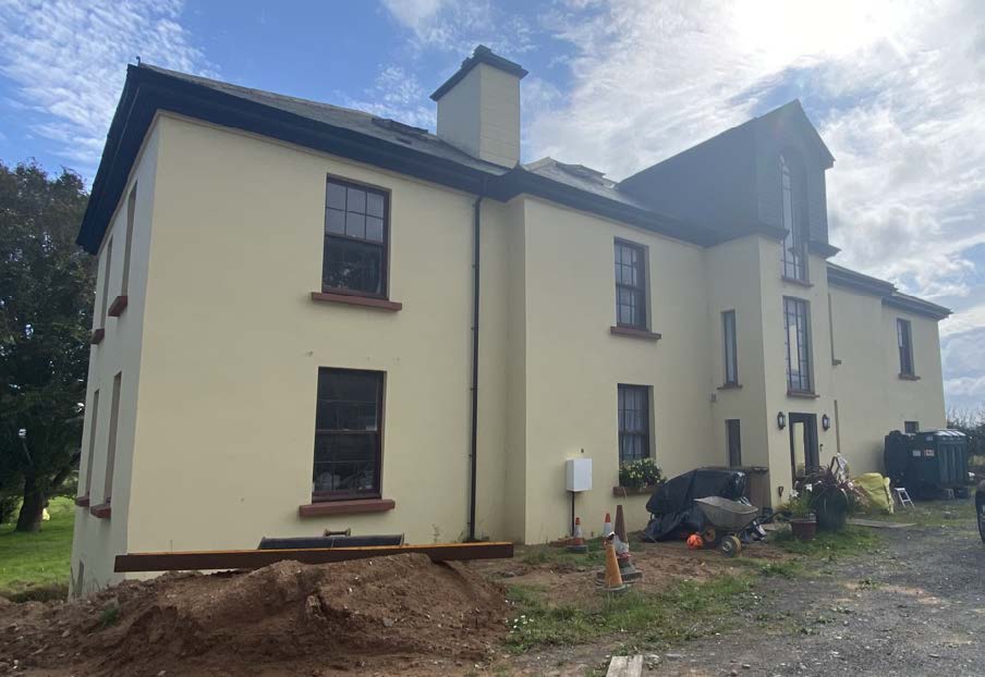 A photograph of a large, two-story cream-colored house with dark window frames, showing construction activity and a pile of earth in the foreground.