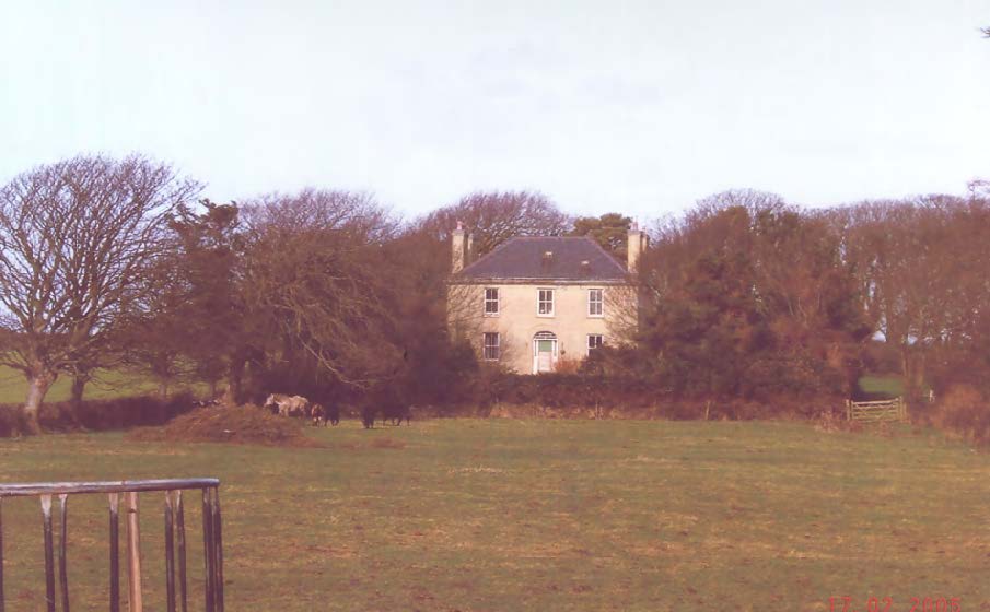 A photograph showing a large detached stone house, Ballaradcliffe House, situated in a rural landscape with trees and a grassy field in the foreground.