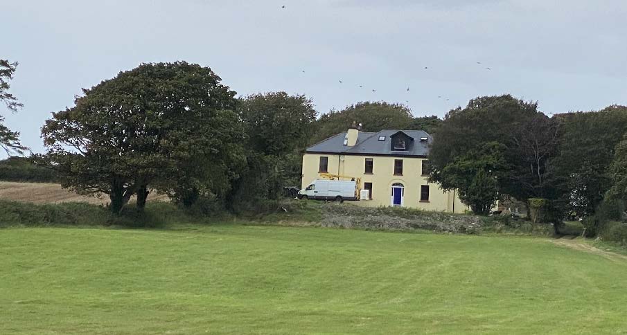 A photograph showing a large, cream-colored detached house situated in a rural setting with trees and a green field in the foreground.