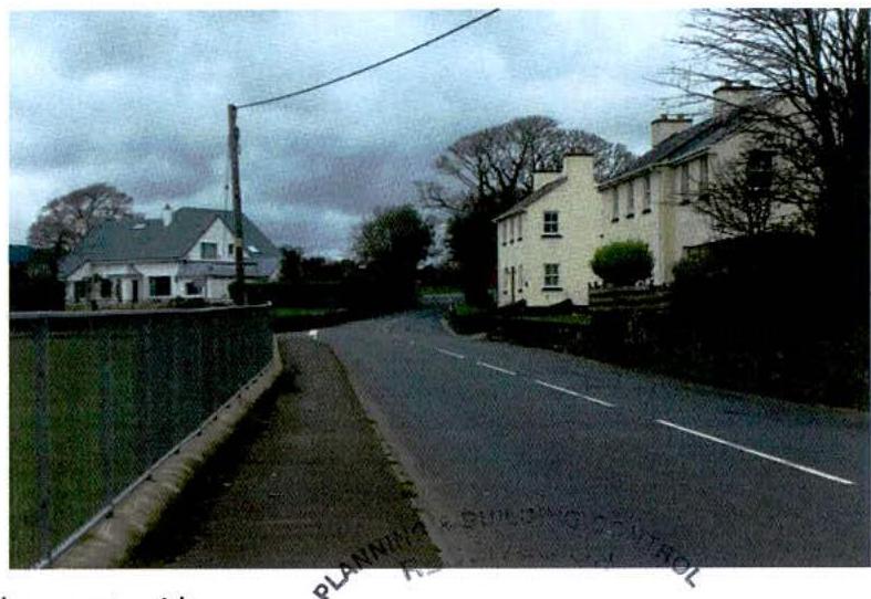 A street-level photograph showing a white two-story house on the right side of a road, with a green fence in the foreground and another house visible in the background.