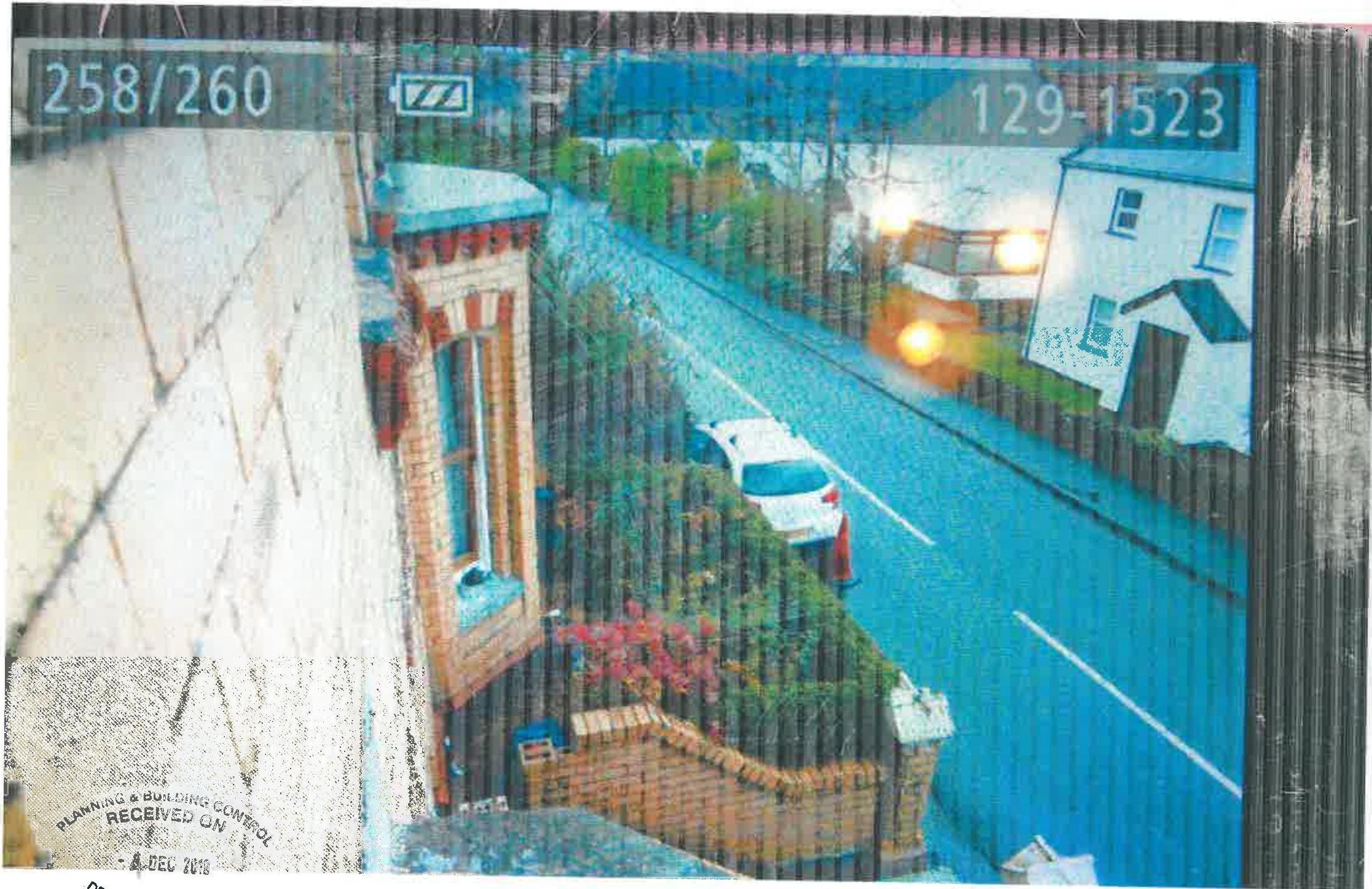 A high-angle photograph showing a residential street scene with a brick house, front garden boundary wall, and a white car parked on the road.