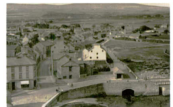 A historical black and white photograph showing a town view with buildings, a road, and a bridge in the foreground, likely depicting the site location or surrounding area.