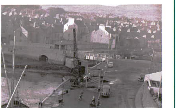 A grainy black and white photograph showing a harbor area with construction equipment in the foreground and a densely built hillside town in the background.