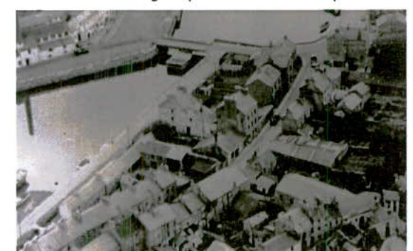 A grainy black and white aerial photograph showing a dense cluster of buildings along a waterfront or harbor area.