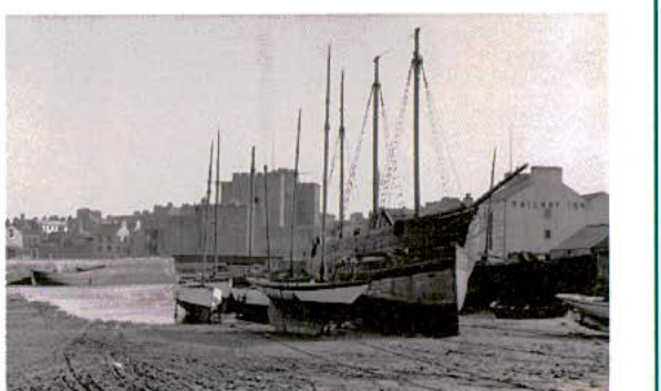 A historical black and white photograph showing several boats resting on a beach or tidal flat with a building labeled 'Railway Inn' in the background.