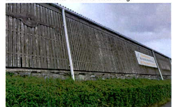 A photograph showing the weathered vertical wooden cladding and stone foundation of a long building with a green hedge in the foreground.