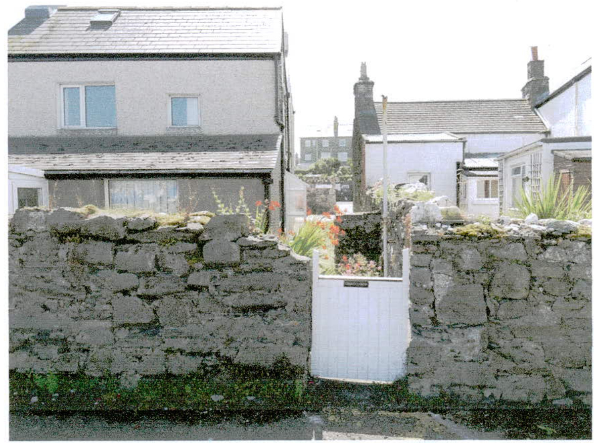 A street-level photograph showing a dry stone wall with a white gate in the foreground, behind which are residential houses with slate roofs.
