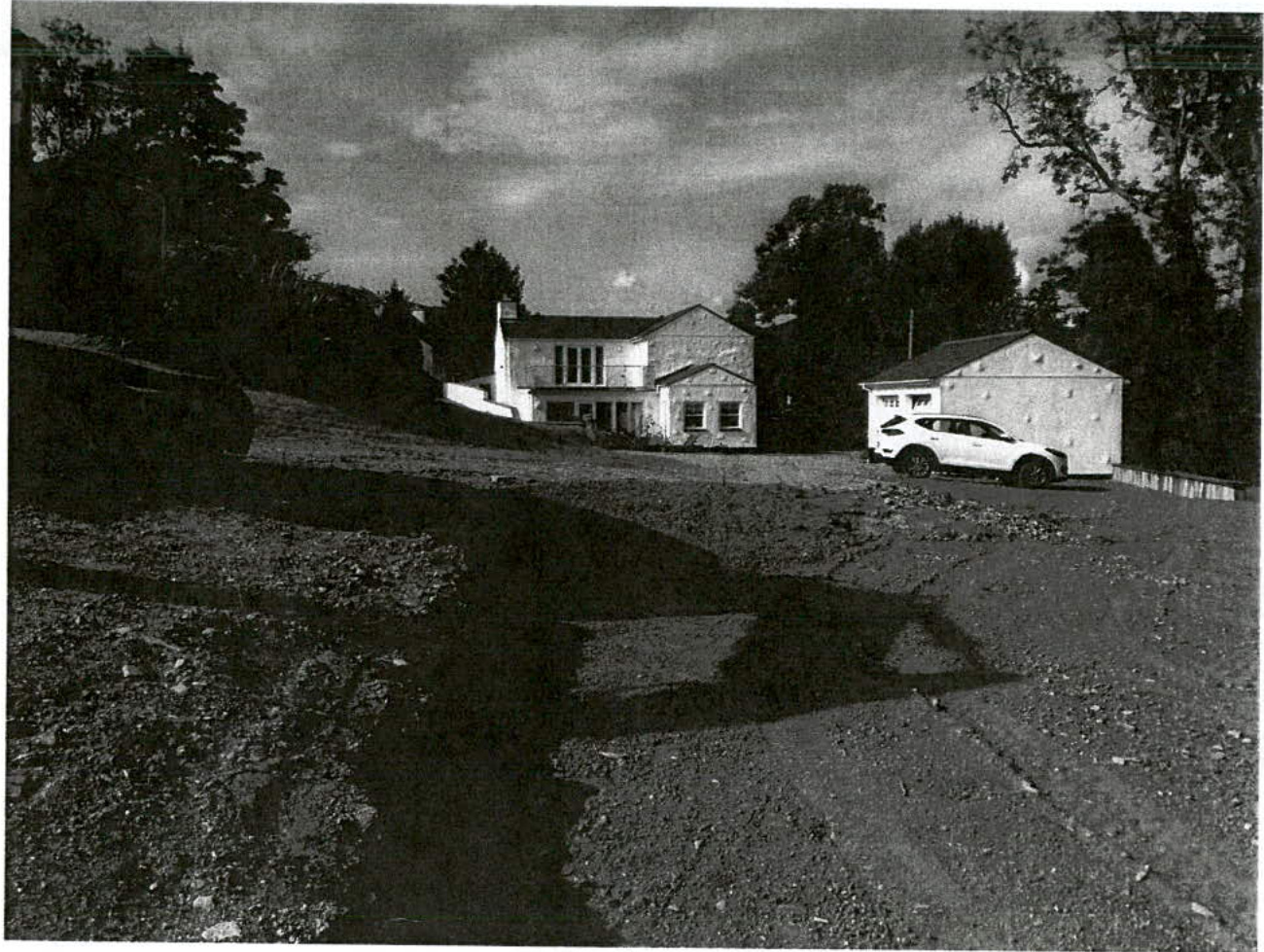 A black and white photograph showing a detached house and garage with a car parked outside, featuring rough, disturbed ground in the foreground consistent with landscaping works.