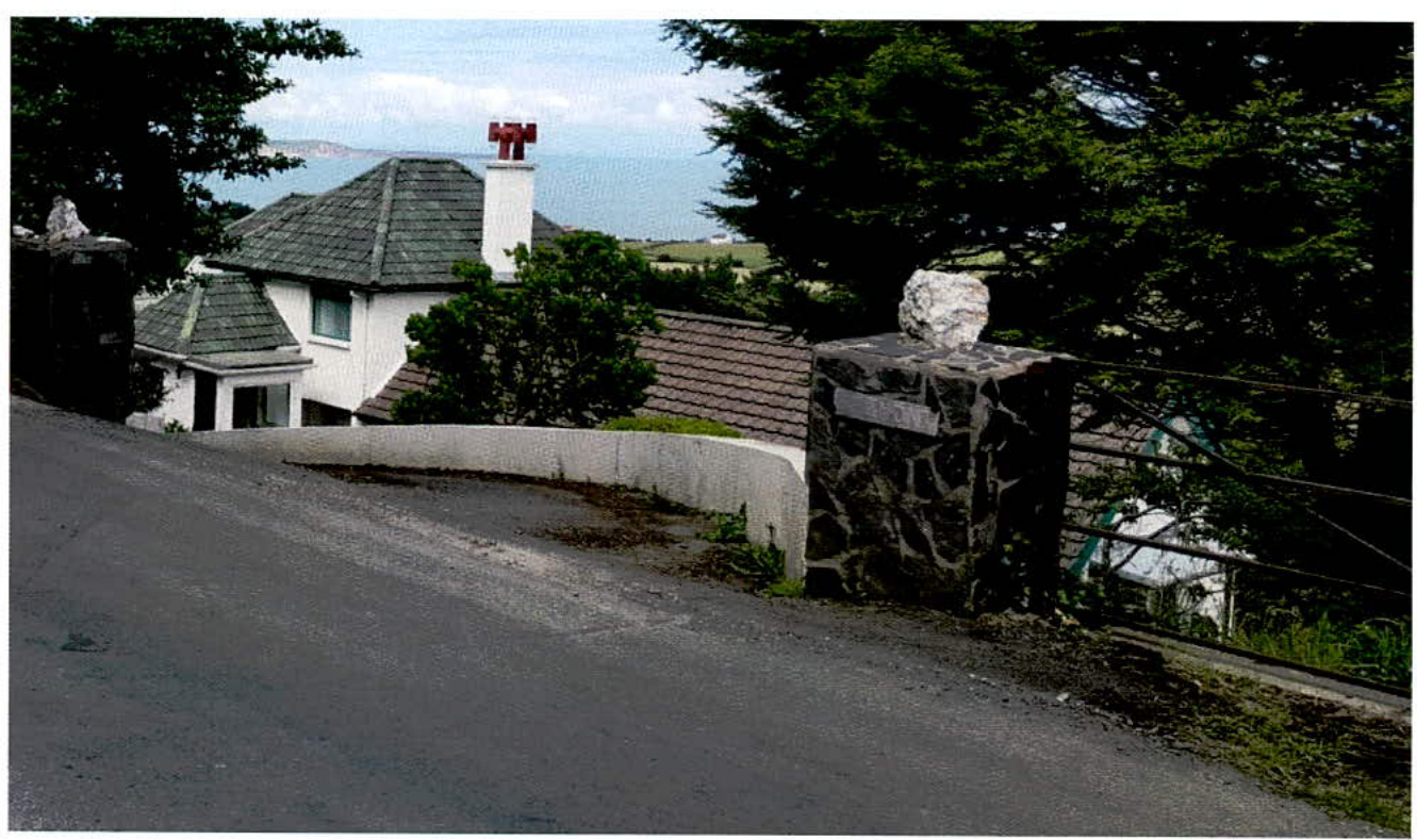 A photograph showing the existing white detached house viewed from the roadside, featuring a stone gate pillar and a coastal background.