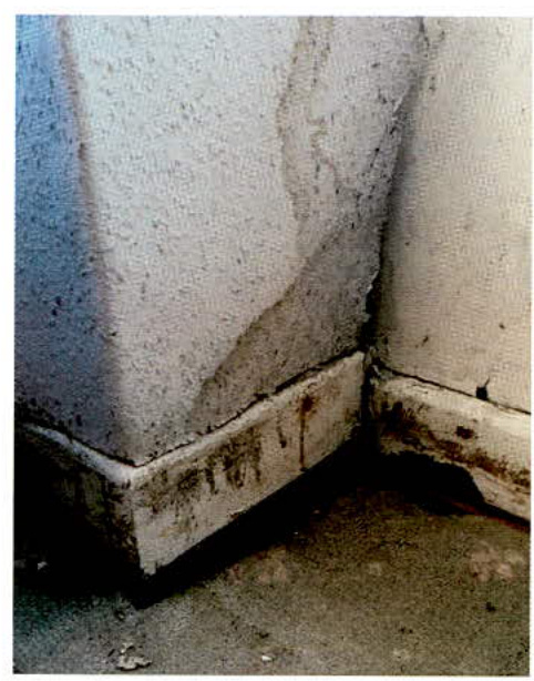 A close-up photograph showing the corner of a building's exterior wall and concrete plinth with visible water staining and dampness.