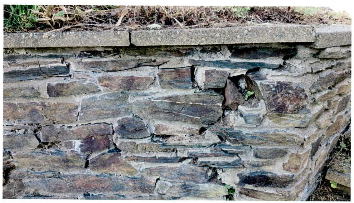 A close-up photograph of an existing stone wall, likely a boundary or retaining wall, constructed from layered slate-like stones with a concrete coping on top.