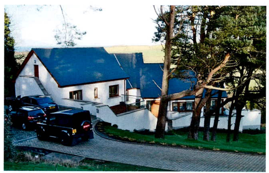 A photograph showing a large white detached house with a dark roof, featuring a paved driveway with parked vehicles and surrounding trees.