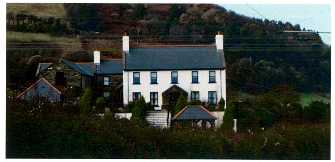 A photograph showing a large white two-story detached house with a slate roof, situated on a hillside with surrounding vegetation and outbuildings.