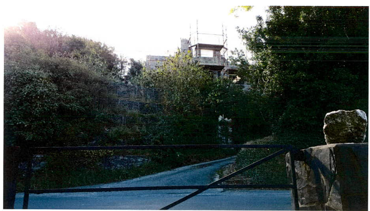 A photograph showing a building under construction on a wooded hillside, viewed from a driveway with a gate in the foreground.
