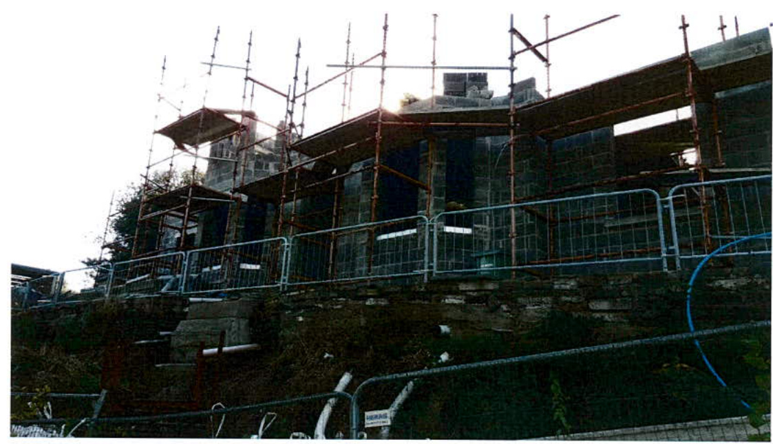 A photograph showing a building under construction with extensive scaffolding and temporary site fencing. The structure features grey blockwork walls and sits behind a stone retaining wall.
