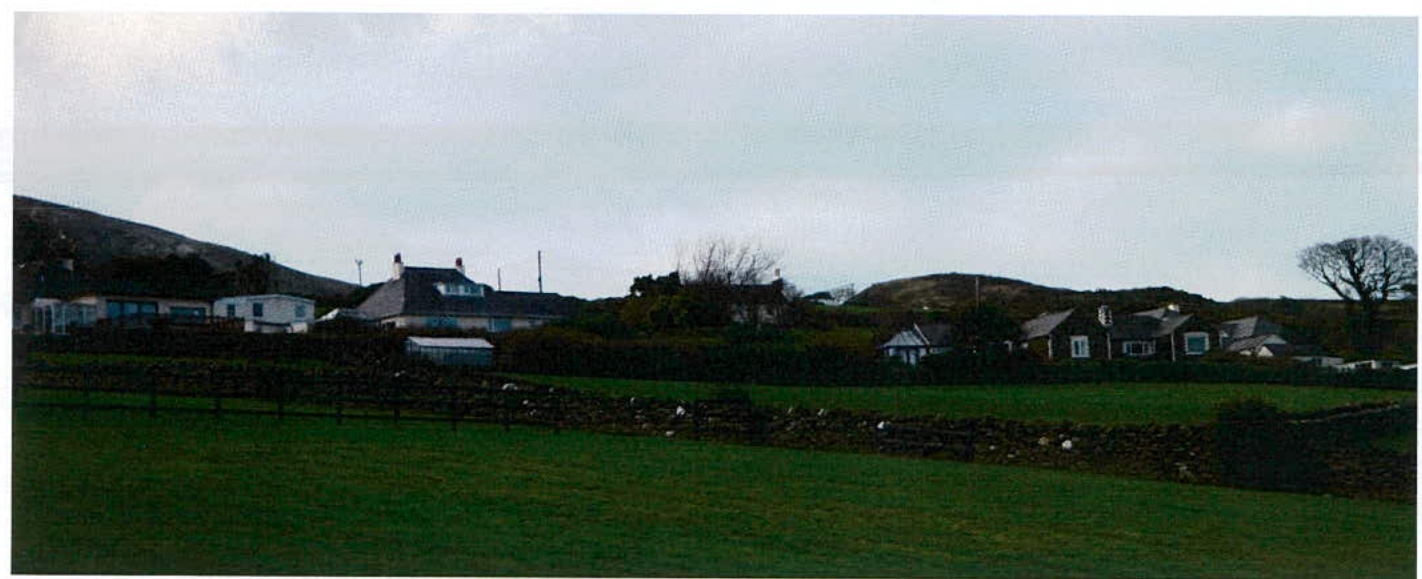 A wide landscape photograph showing a rural scene with a green field in the foreground, a dry stone wall, and several houses situated on a hillside in the background.