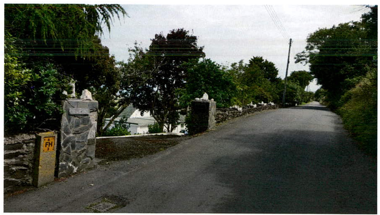 A street-level photograph showing a paved road curving uphill, bordered by a stone wall and gateposts on the left with a white house visible behind the trees.