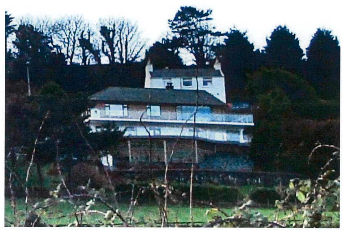 A grainy photograph showing a large, multi-story white detached house situated on a sloping site with trees in the background.
