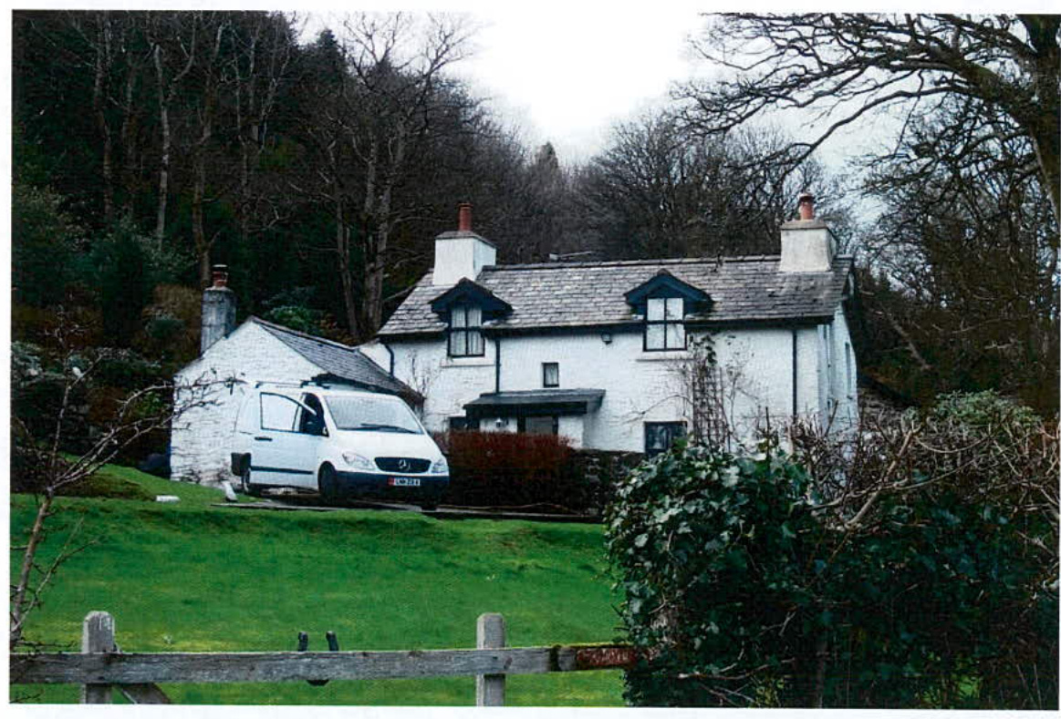 A photograph showing a white, two-story detached house with a slate roof situated on a grassy slope with a wooded background.