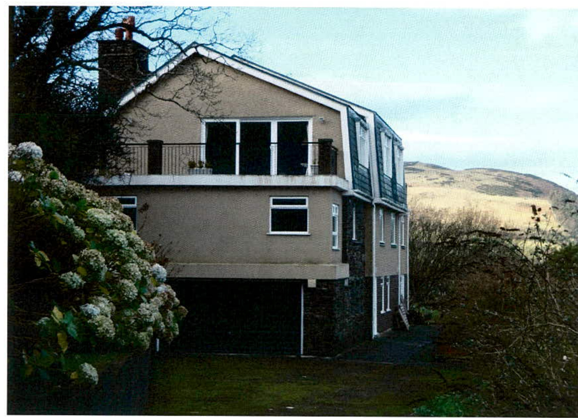 A photograph of a two-story detached house featuring a balcony and garage area, situated in a rural setting with hills in the background.
