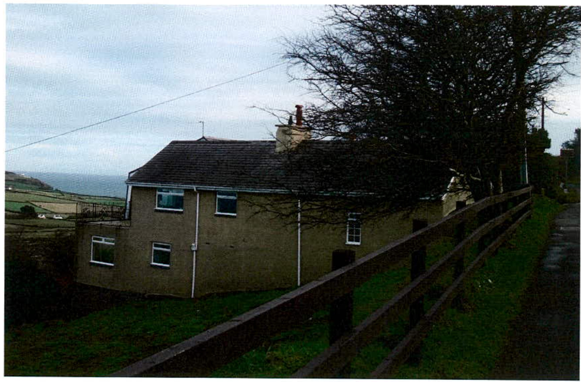 A photograph showing an existing two-story detached house with a slate roof, situated on a slope with a coastal view in the background.