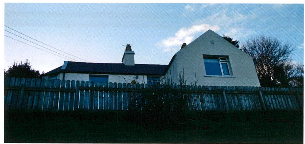 A photograph of a white, single-story detached bungalow situated behind a wooden fence in a rural setting.