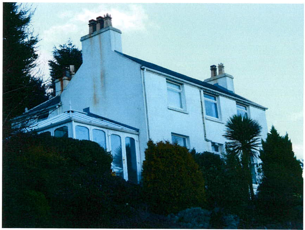 A grainy photograph showing the exterior of a white, two-story detached house with a conservatory extension, surrounded by mature trees and shrubbery.