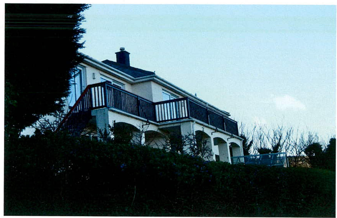 A photograph of a two-story detached house featuring a prominent upper-level balcony with dark railings and arched openings on the ground floor. The property is situated on a slope with dense vegetation and trees surr...