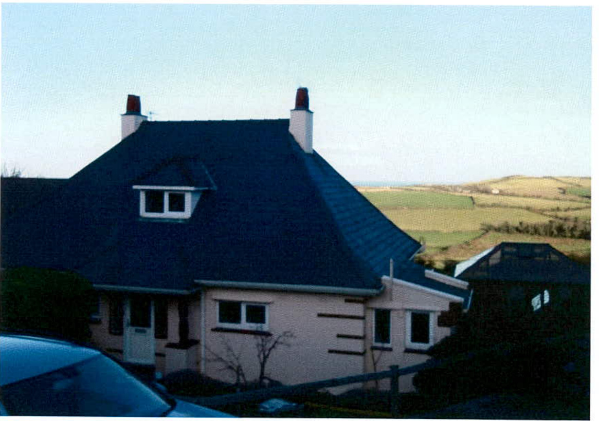 A photograph showing a detached house with a dark roof and white chimneys, situated in a rural setting with fields and a distant horizon visible in the background.