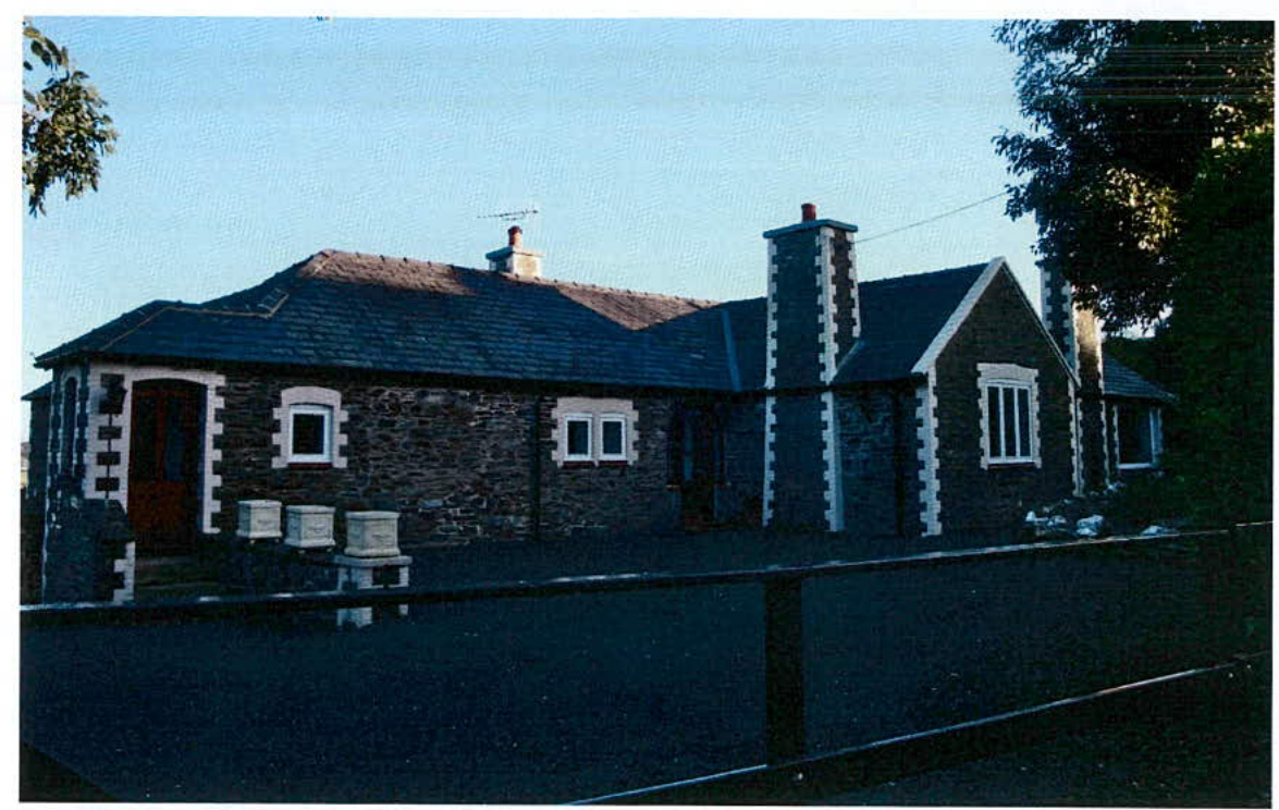 A photograph of an existing stone detached bungalow with white quoins and window frames, situated near a road.