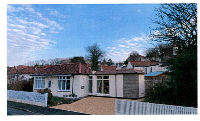 A photograph of a single-story bungalow featuring a side extension with large windows and a tiled roof, viewed from the street with a white fence in the foreground.