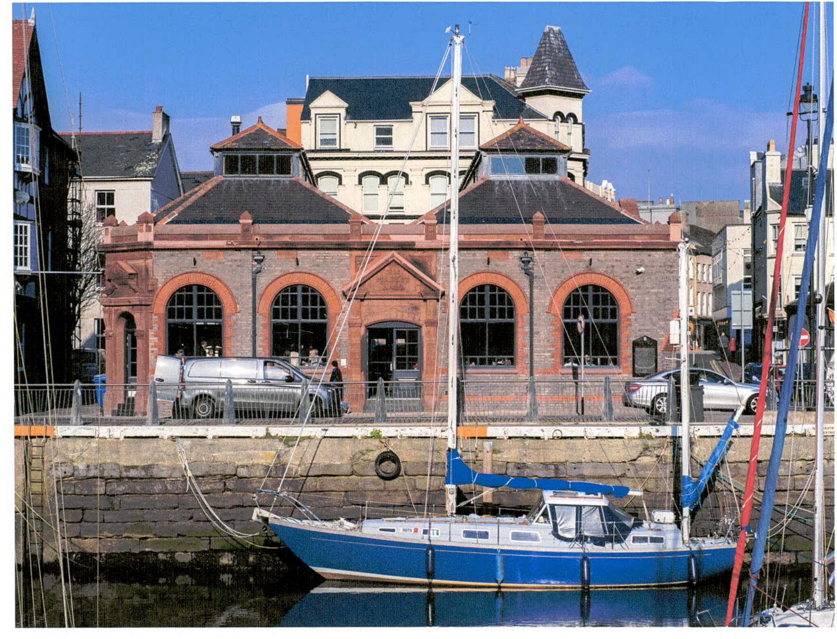A photograph showing a red brick building with arched windows situated on a harbor quay, with a blue sailboat in the foreground.