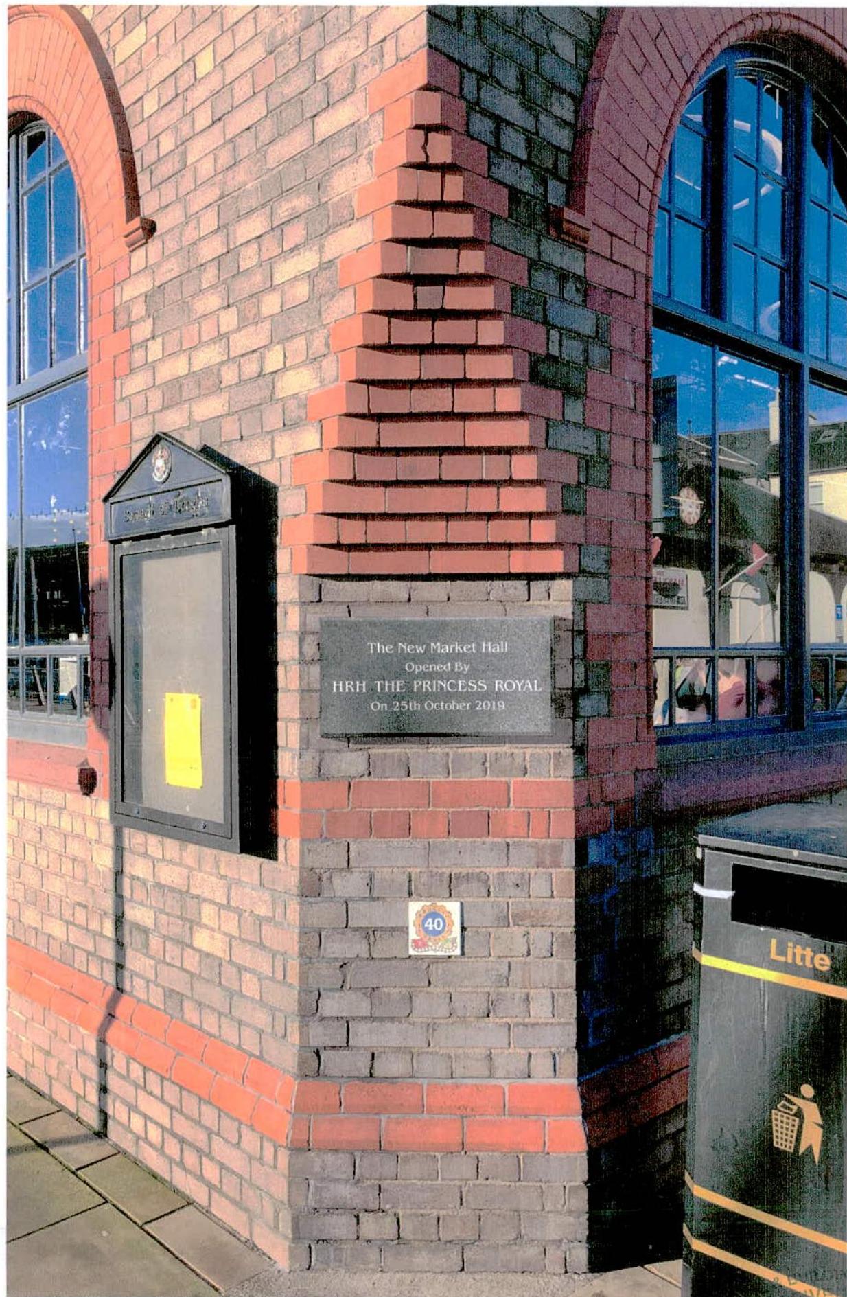 A photograph showing the exterior brick corner of a building, featuring a mounted granite commemorative plaque and a notice board.