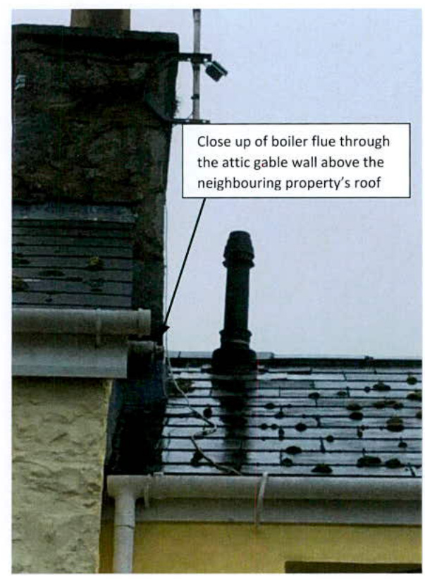 A close-up photograph showing a black boiler flue pipe protruding from a slate roof next to a stone gable wall, accompanied by an annotation explaining the flue's position relative to a neighboring property.