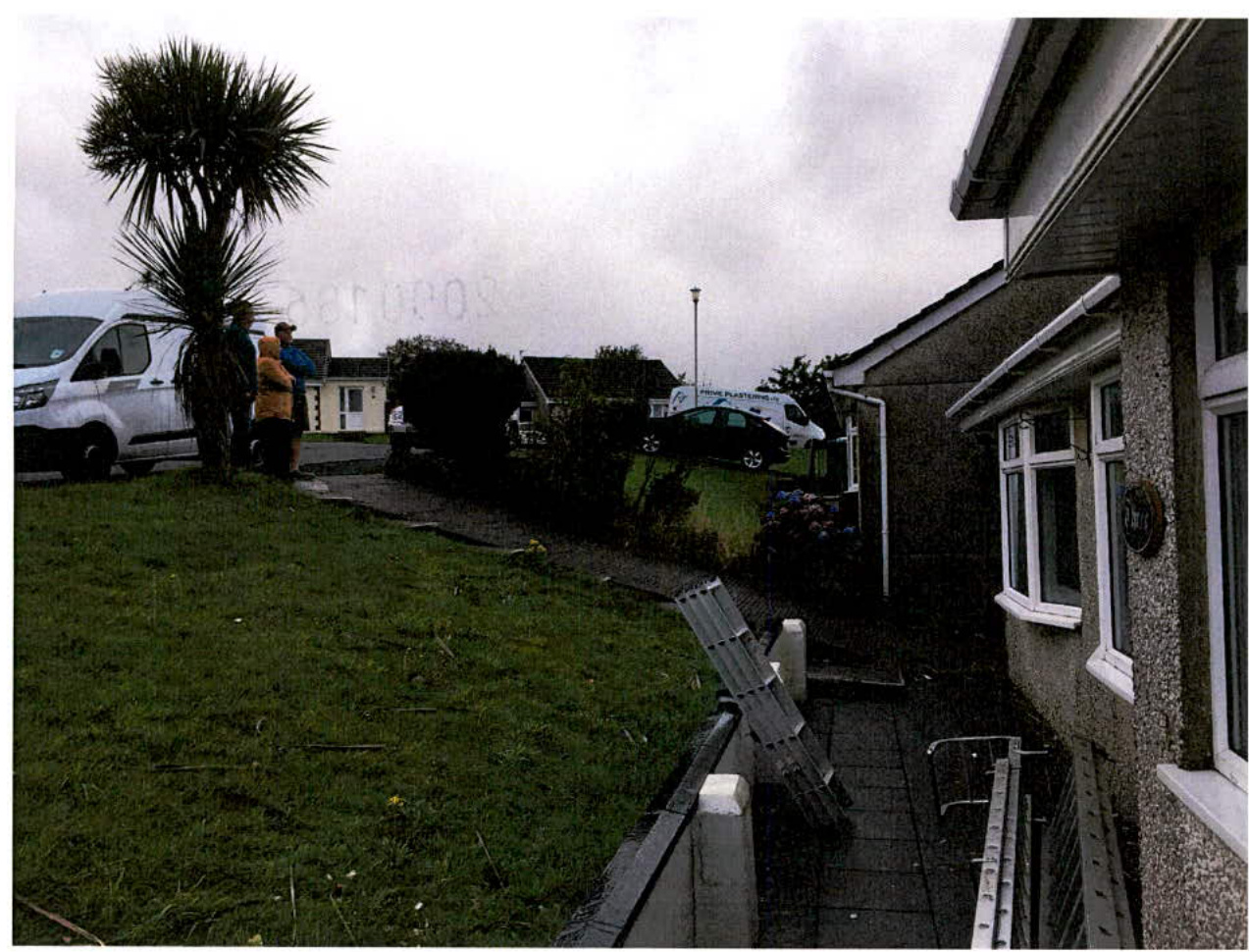 A photograph showing the sloping front garden and driveway area of a residential property, with a house on the right and vehicles parked on the street to the left.