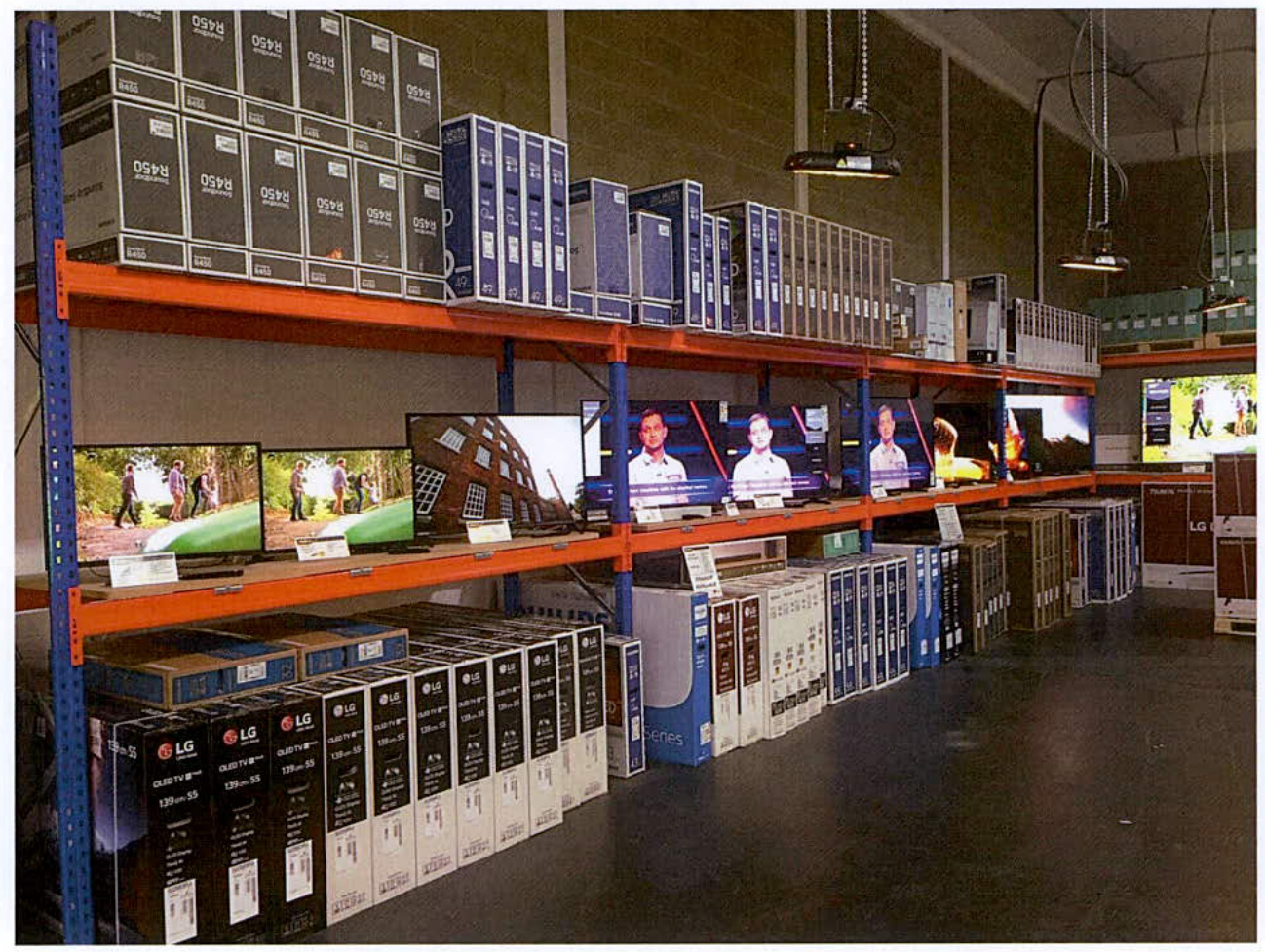 This photograph shows the interior of a commercial warehouse or storage facility. Industrial shelving units are stocked with boxes of electronics, including OSRAM lighting and LG televisions, with several screens disp...