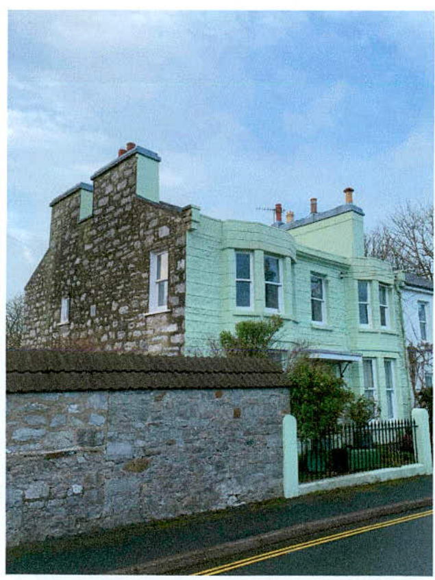 A street-level photograph showing a building facade with a mix of exposed stone and painted render, situated behind a stone boundary wall. The image visually documents the current state of the front elevation relevant...