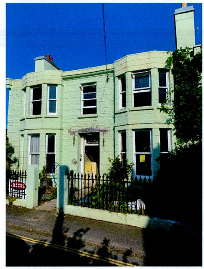 A photograph showing the front elevation of a two-story, pale green terraced house with bay windows and a black metal boundary fence.