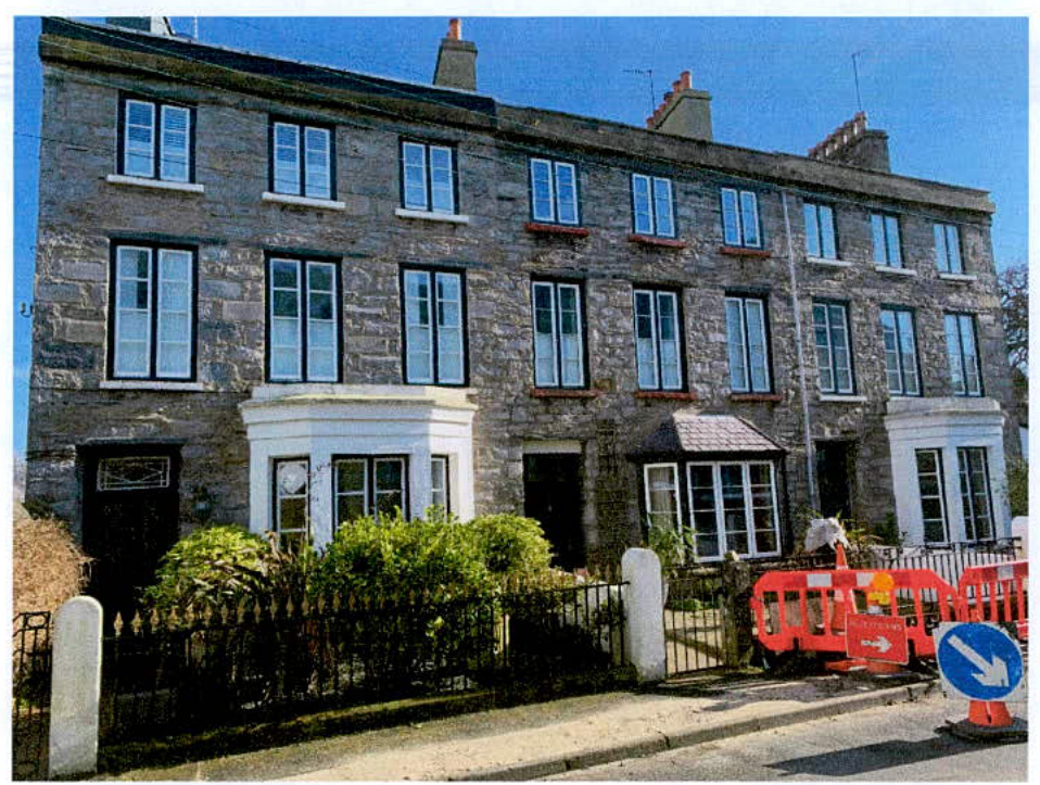 A street-level photograph of a long stone terraced building with exposed masonry, white bay windows, and red construction barriers in the foreground.